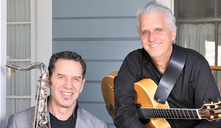 2 men, one with a saxophone and one with a guitar in front of a wall